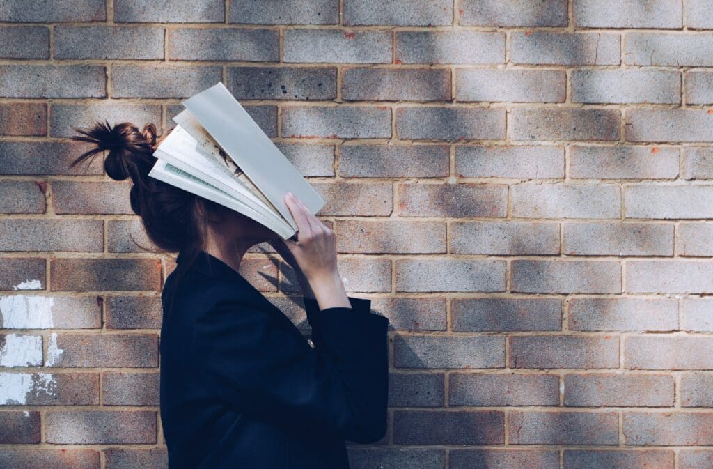 Young adult girl covers her face with a book out of emarassment. She's standing with a side profile in front of a brick wall.