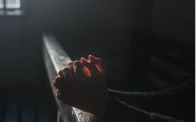 The folded hands of someone kneeling in prayer are resting on the top of a church pew in a dimly lit sanctuary.