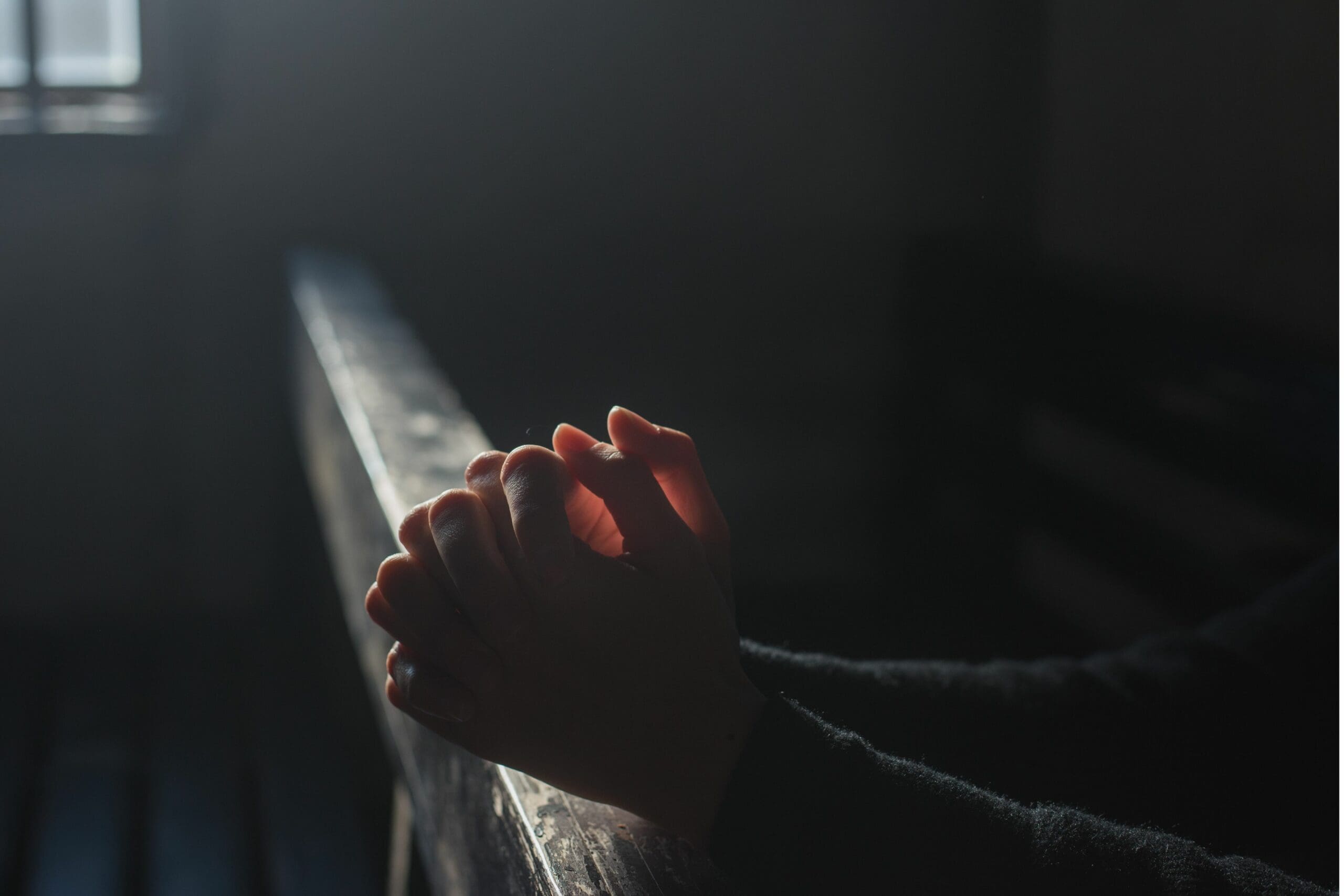 The folded hands of someone kneeling in prayer are resting on the top of a church pew in a dimly lit sanctuary.