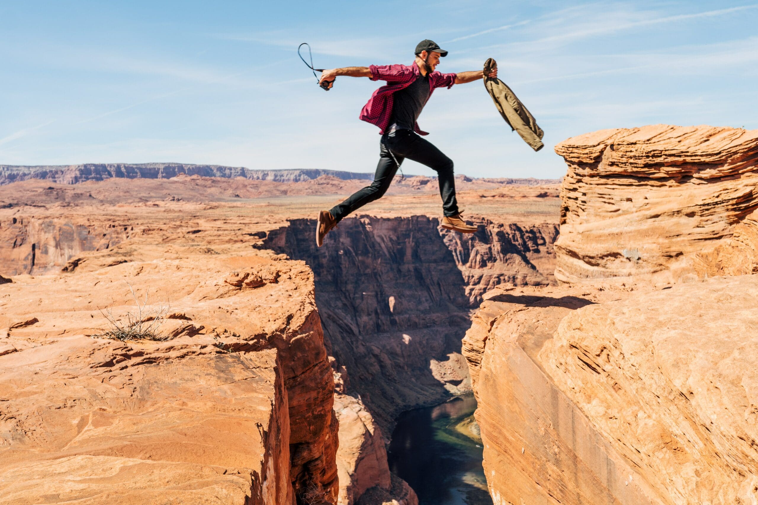 A man jumping across a canyon suspended mid-air. He's carrying a camera in one hand and a jacket in the other. The depth of the canyon is clear below and a river is running through it.