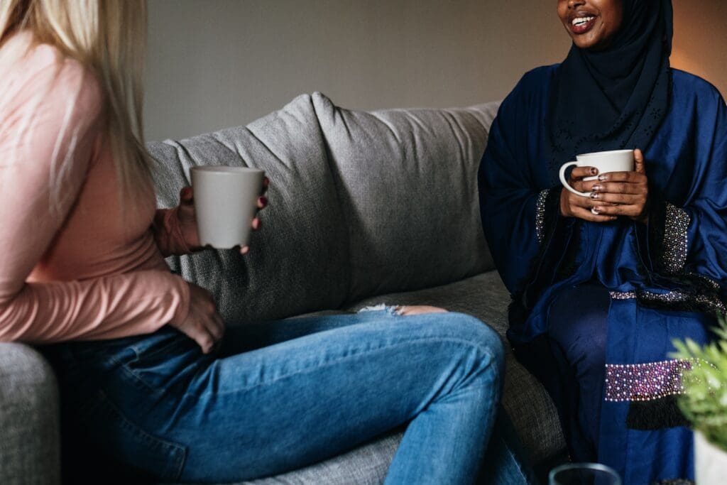 An american womann and a foreign woman are sitting together on a couch in a living room. They are enjoying conversation over a hot beverage.