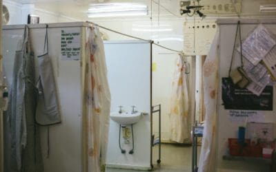 A single room of a developing world hospital. thin, temporary wall dividers separate each hospital bed and the fourth wall is a plastic shower curtain. Charts, aprons and hand washing sinks hang from the walls. Bright flourescent lights glare off the ceiling.