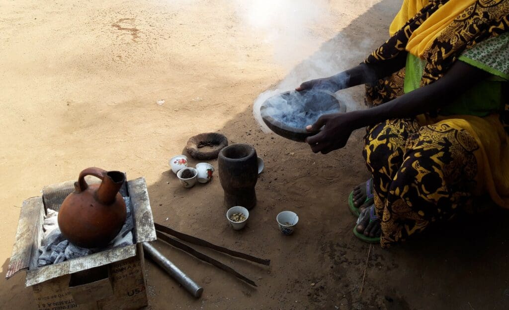 A North African in the middle of a ceremonial coffee ritual. Smoke can be seen wafting from the hot coals in the bowl as she holds them. Cups, a mortar and pestle, and all the tools needed to make coffee can be see on the ground.