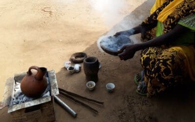 A North African in the middle of a ceremonial coffee ritual. Smoke can be seen wafting from the hot coals in the bowl as she holds them. Cups, a mortar and pestle, and all the tools needed to make coffee can be see on the ground.