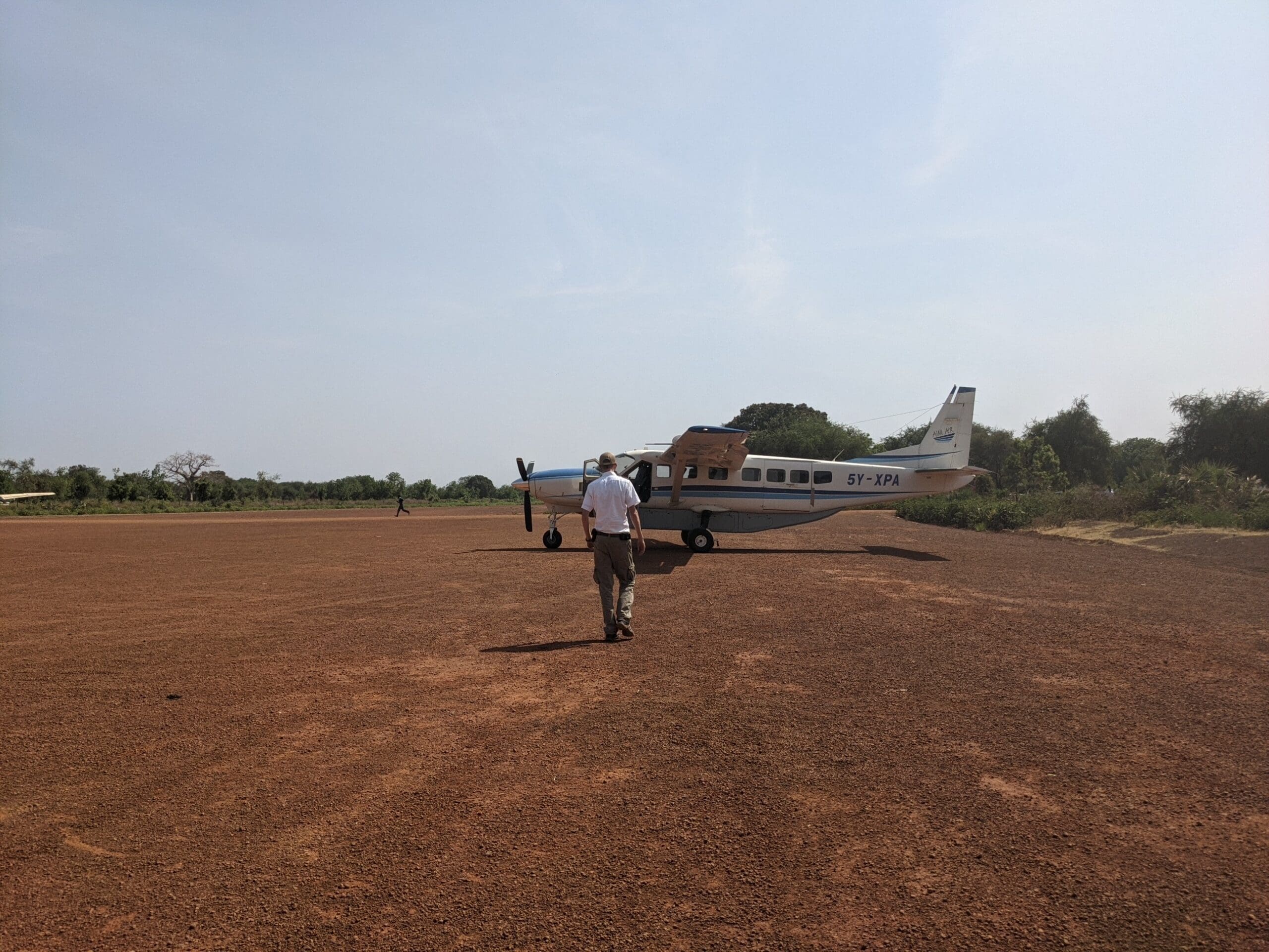An East African pilot walks along the red dirt runway to a white, blue-striped, single prop plane.