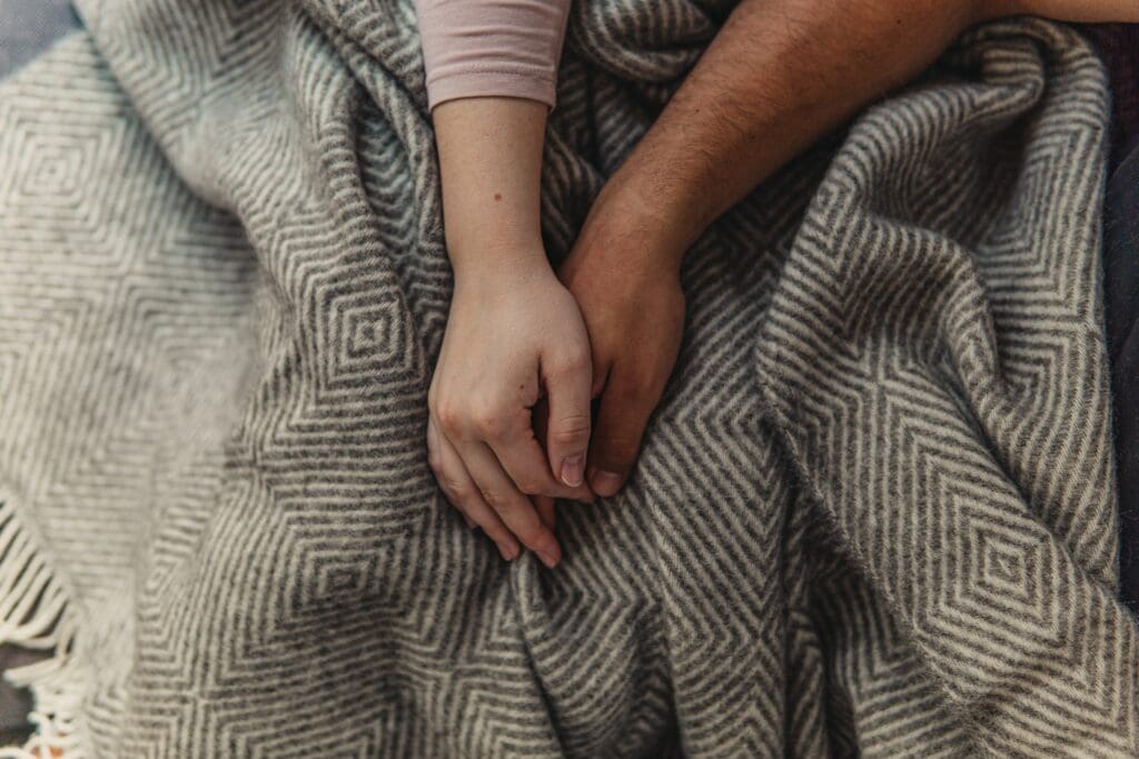 A husband and wife loosely holding hands over the covers.