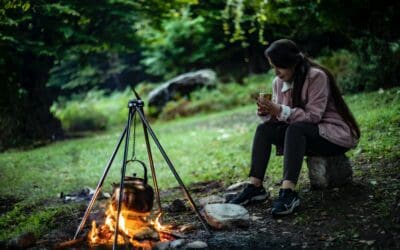 A woman dressed for a hike sits in the middle of a clearing. She is nursing a mug of tea as a kettle warms over the campfire.