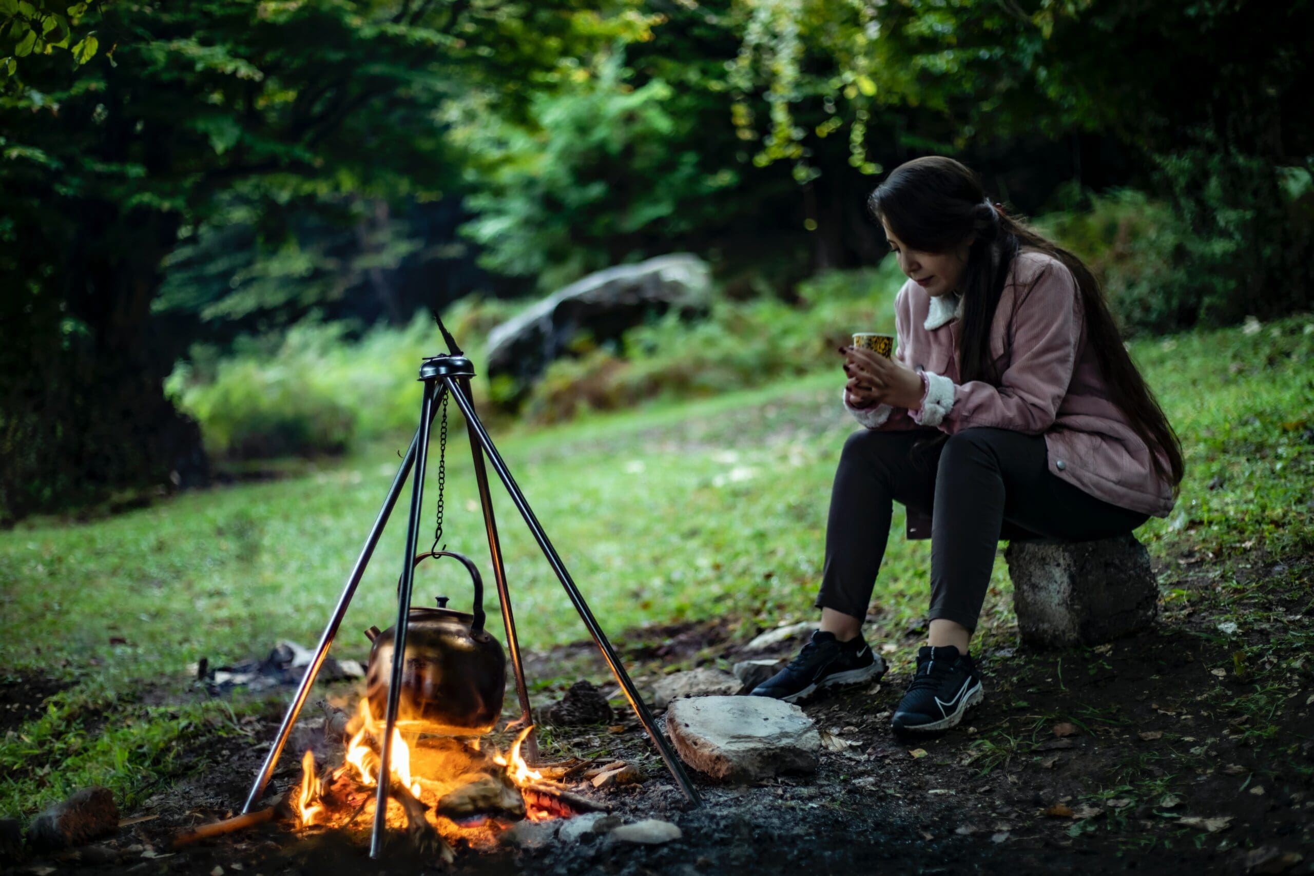 A woman dressed for a hike sits in the middle of a clearing. She is nursing a mug of tea as a kettle warms over the campfire.