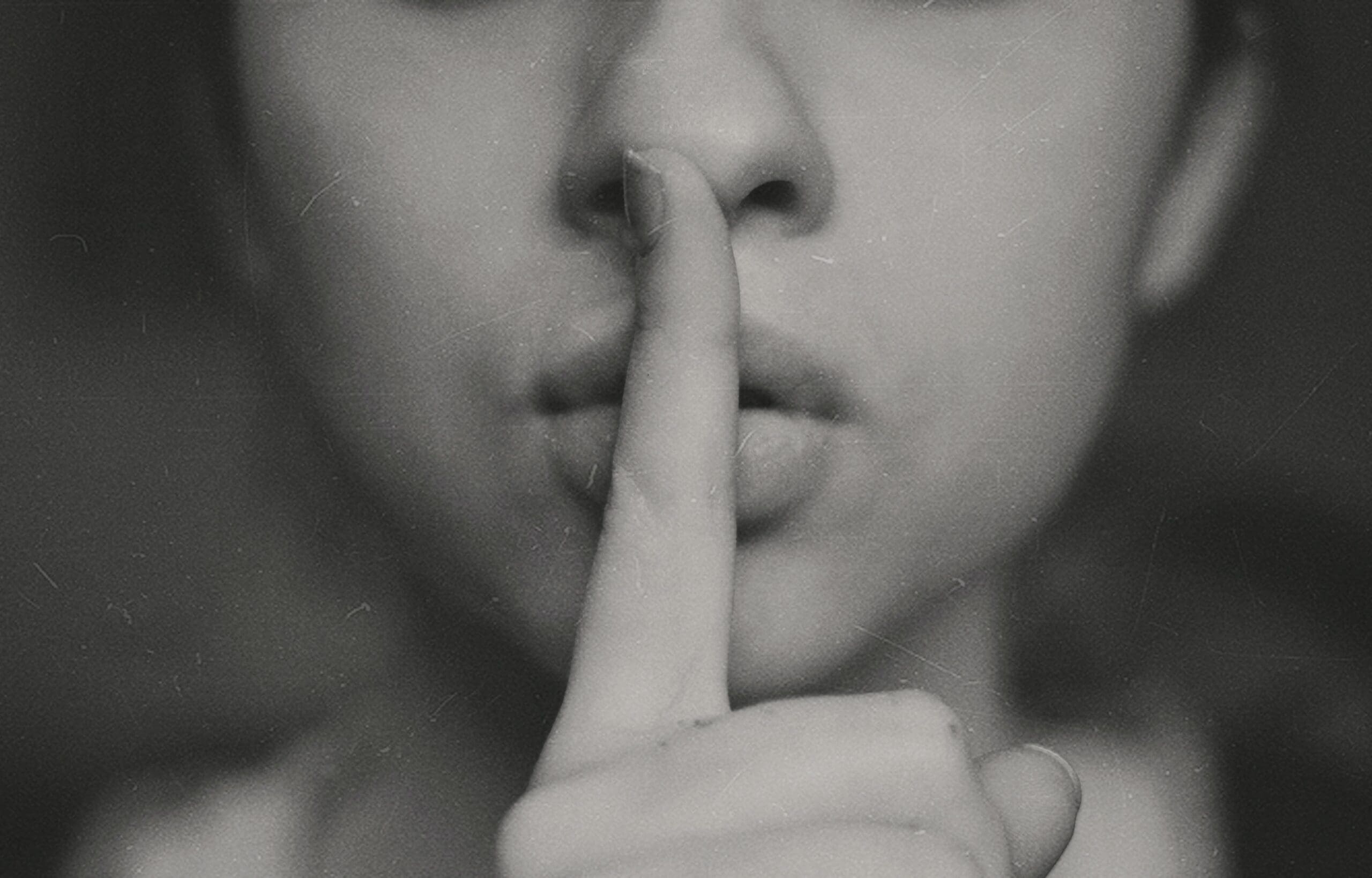A close up portrait from the nose down of a young woman with her pointing finger over her lips. She's shushing someone.